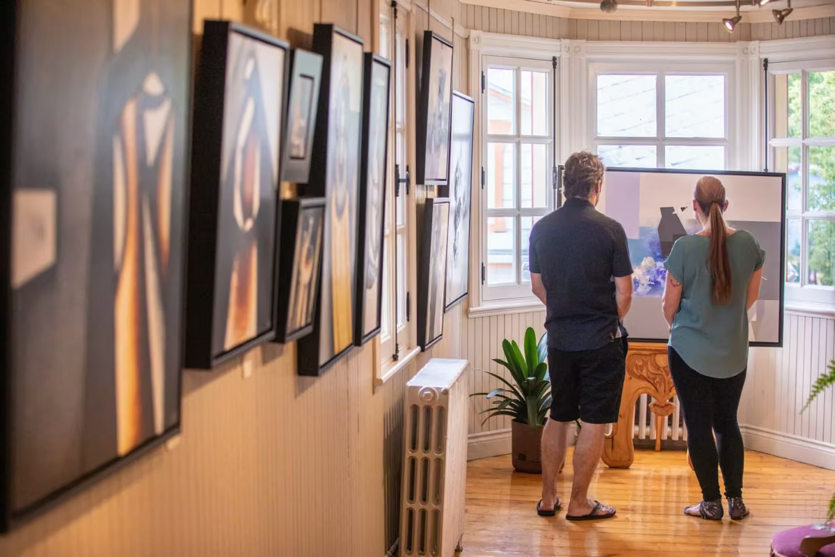 Visiteurs devant des toiles encadrées dans une galerie d'art de Charlevoix, éclairage chaleureux (attrait régional de Charlevoix)