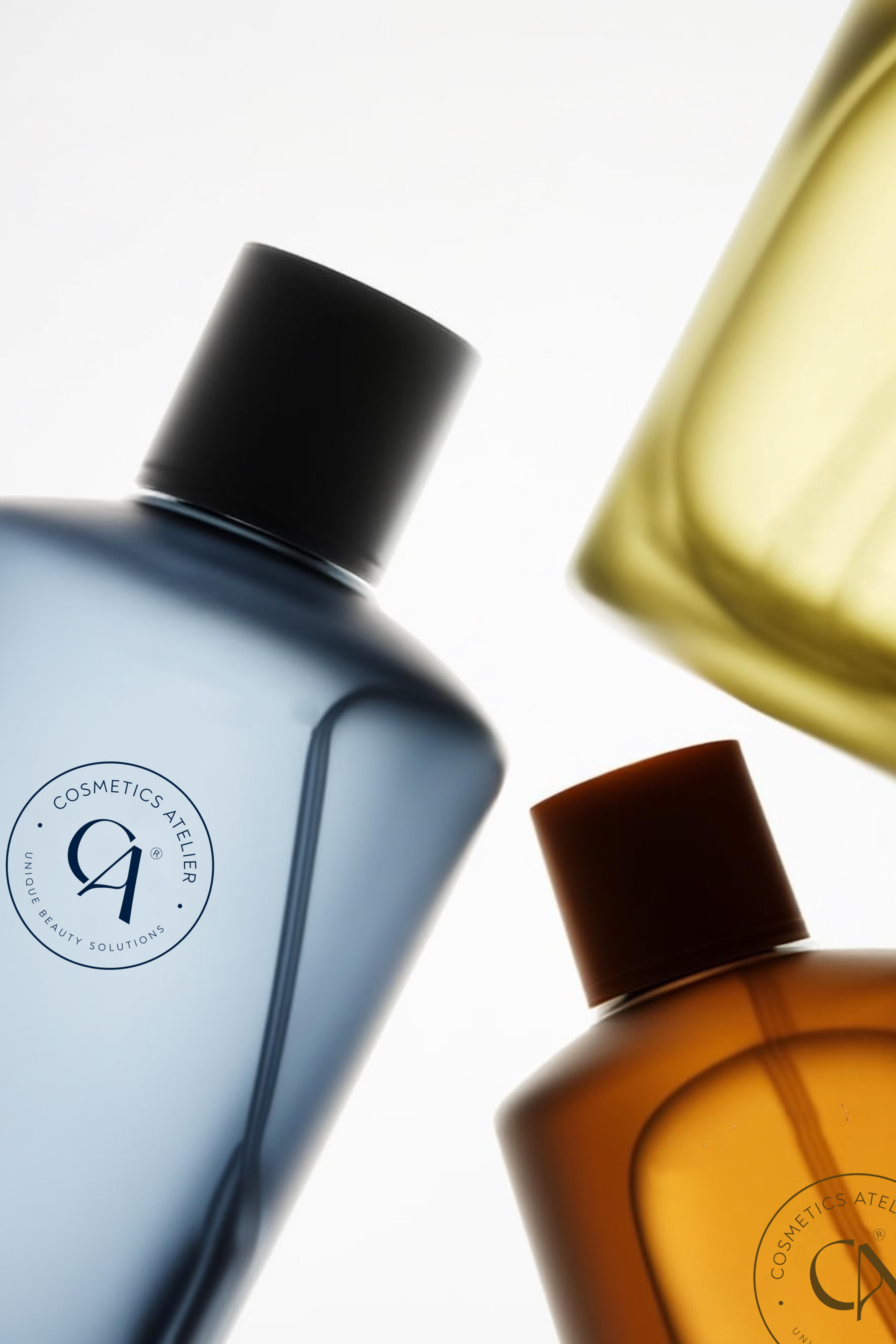 Close-up of three translucent cosmetic bottles in blue, amber, and yellow with black and brown caps, showing partial logos reading Cosmetics Atelier Unique Beauty Solutions on the bottles.