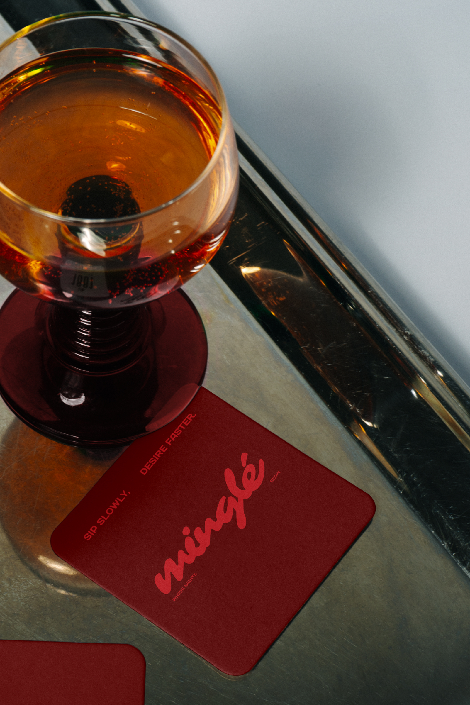 Close-up of a glass with amber liquid on a reflective metal surface next to a dark red coaster with the word 'miingle' in cursive and text reading 'sip slowly. desire faster.'