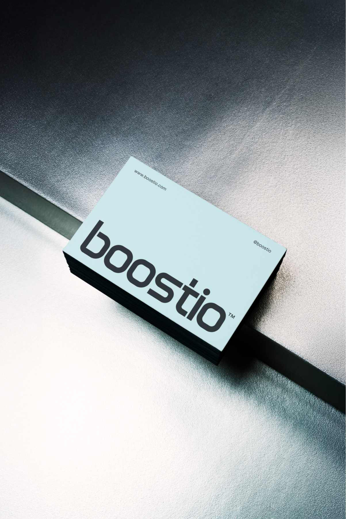 Stack of light blue business cards on a textured metallic surface, showing the brand name 'boostio' in large black letters, with the website www.boostio.com and the handle @boostio printed in small text.
