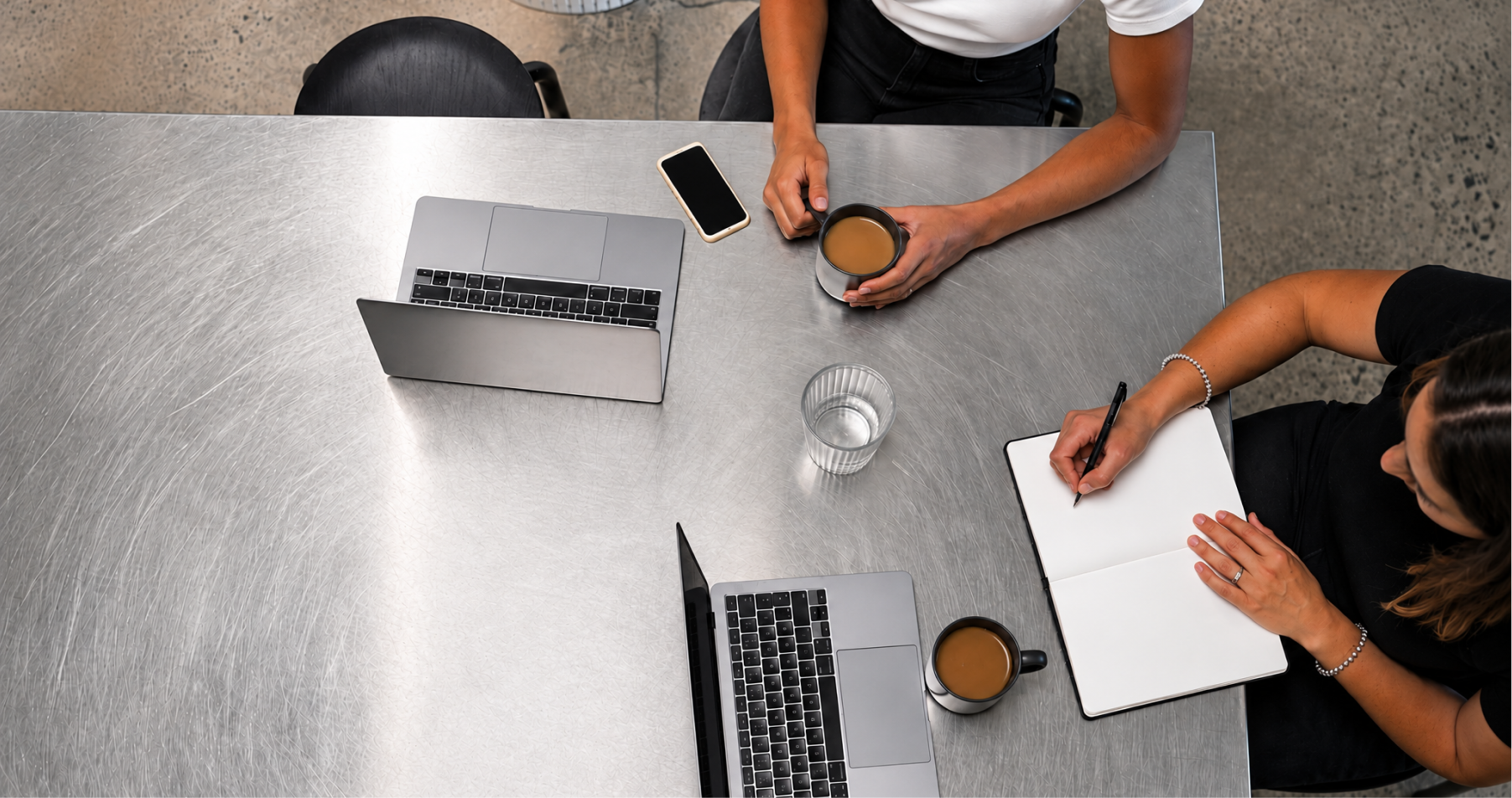 Overhead view of two people working at a metal table with laptops, coffee mugs, a smartphone, a glass of water, and one person writing in a notebook.