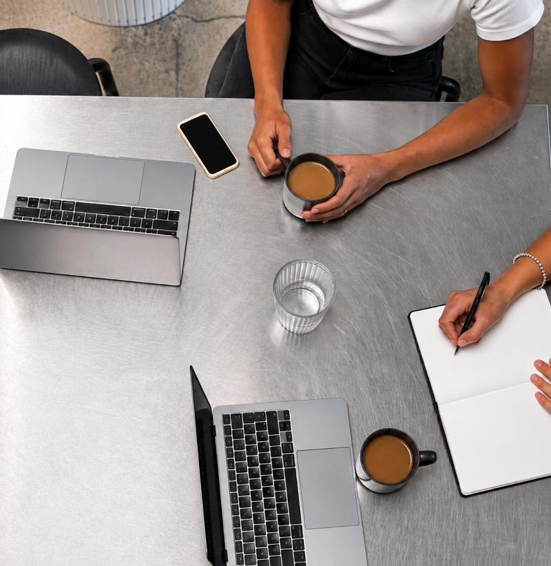 Top view of two people sitting at a metal table with two laptops, a notebook where one person is writing, two cups of coffee, a glass of water, and a smartphone.