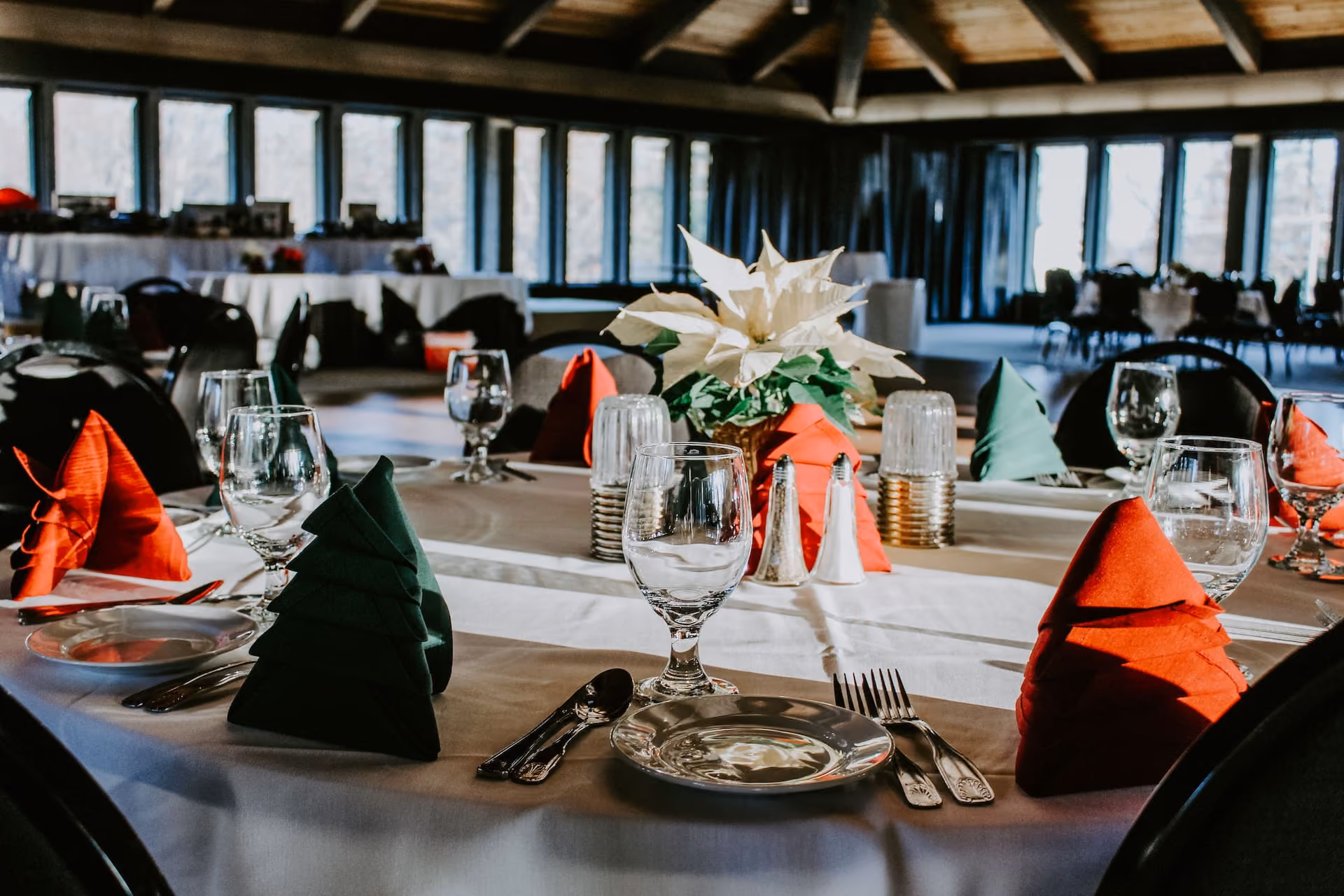 Table set for dining with folded red and green napkins, silverware, glasses, and a white poinsettia centerpiece.