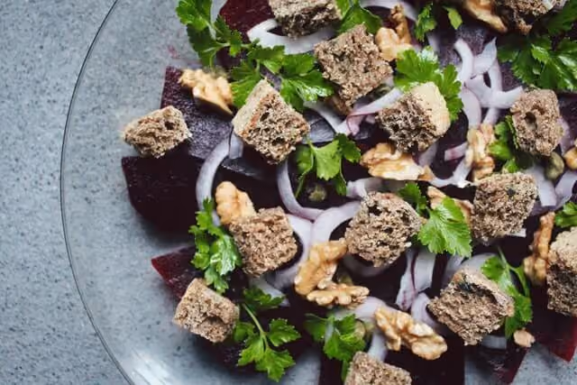 Salad with beetroot slices, walnut halves, red onion slices, parsley leaves, and bread croutons in a glass bowl.