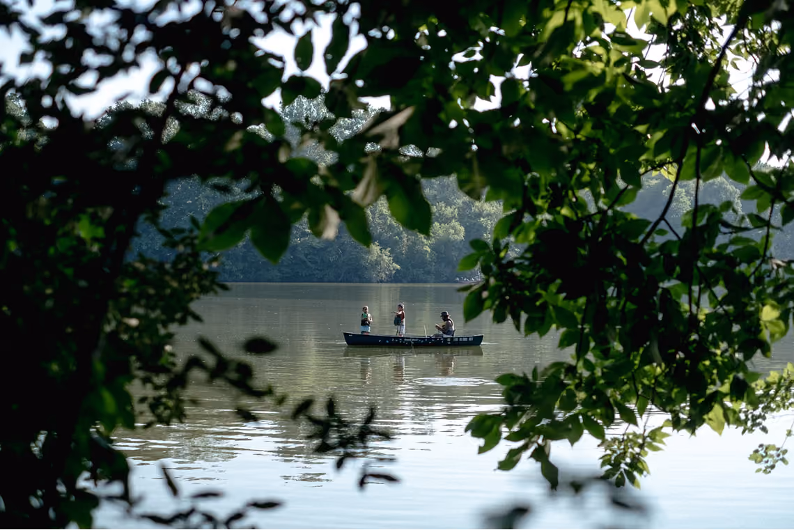 Three people fishing from a boat on a calm lake framed by green tree branches.