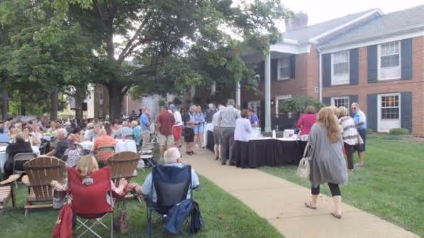 Outdoor social event with people seated and standing near tables on lawn by brick building.