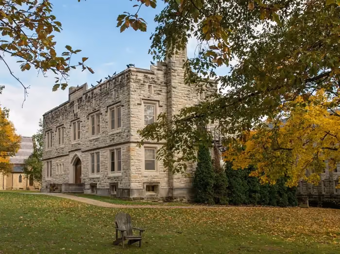 Stone building with arched door, surrounded by trees with autumn leaves and an empty wooden chair on grass.