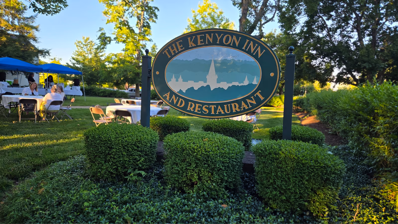 Sign for The Kenyon Inn and Restaurant surrounded by green bushes and outdoor tables with chairs.
