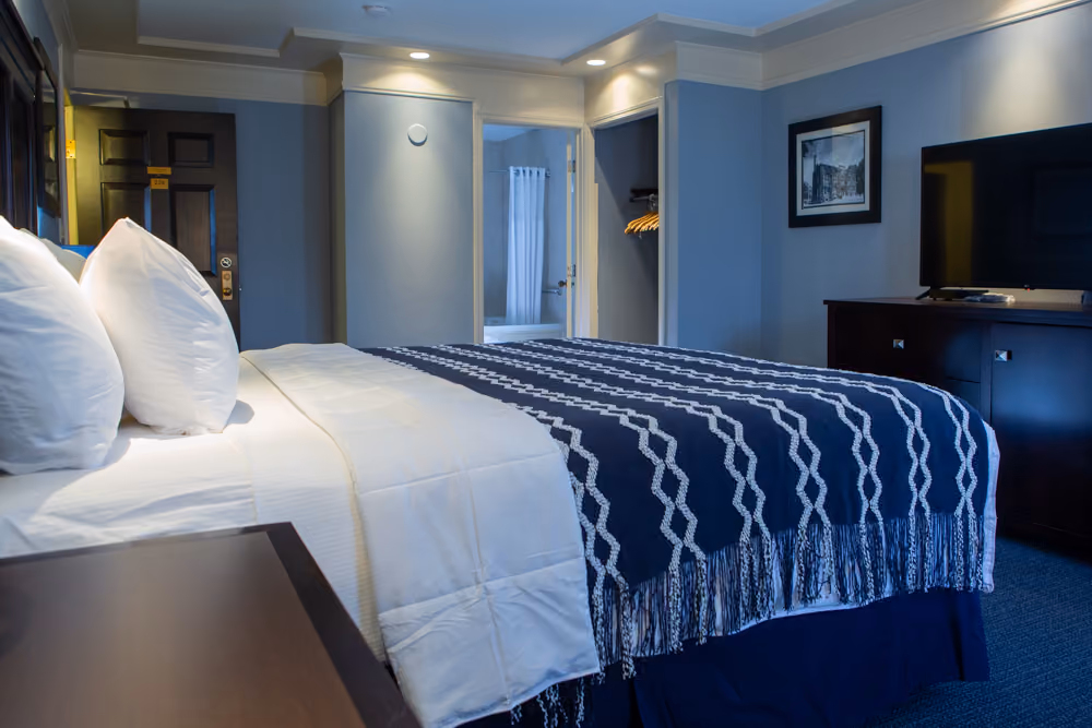 Hotel room with a king bed, white and blue bedding, TV on dresser, open closet, and bathroom doorway.