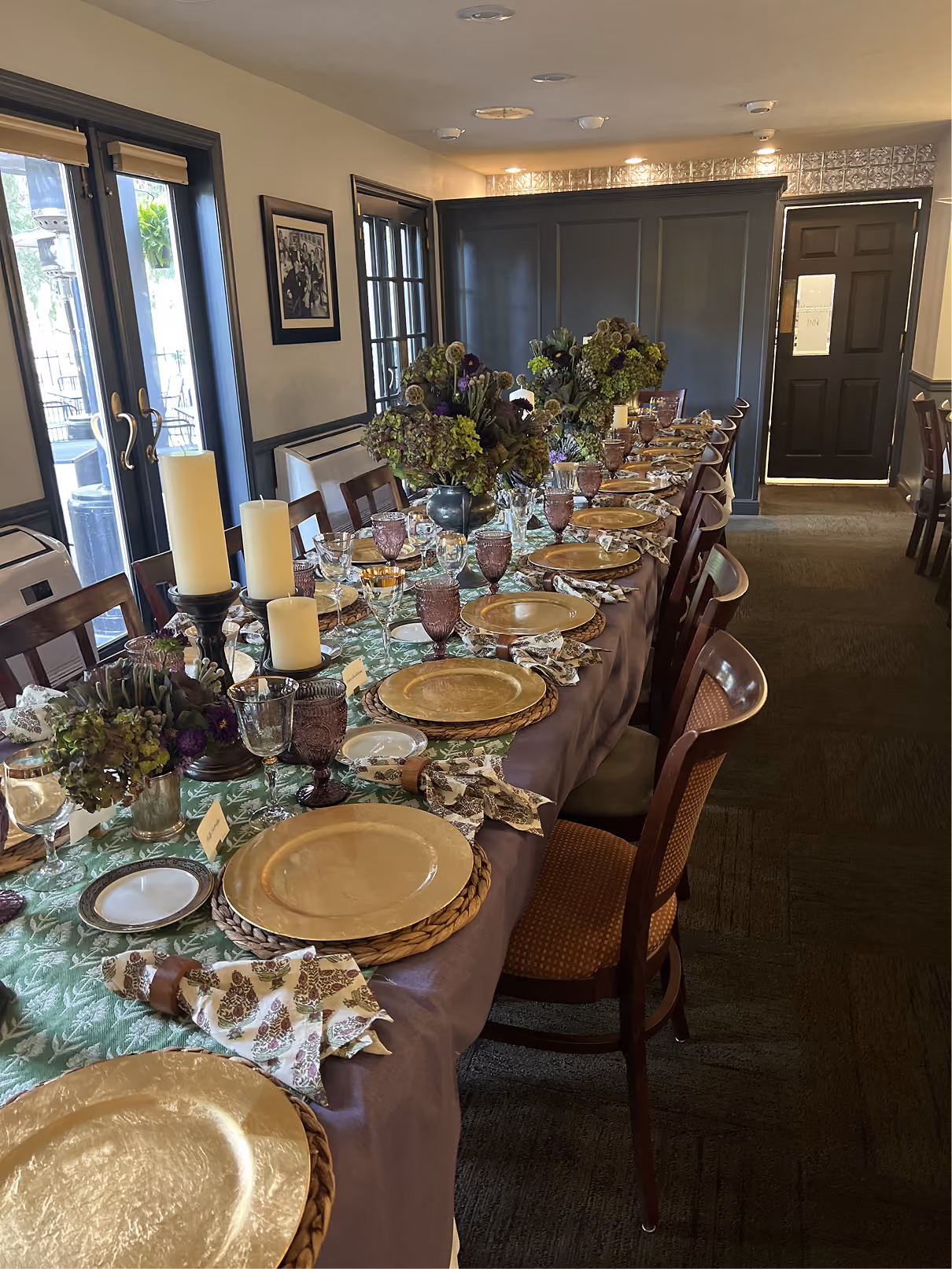 Long dining table set with gold plates, floral napkins, purple glasses, candles, and flower centerpieces in a room.