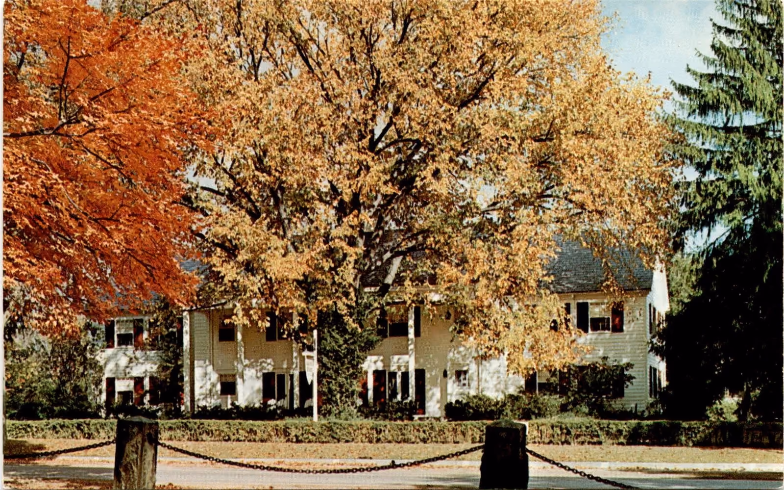 The Alumni House, Gambier, Ohio.
A large white colonial-style house partially obscured by mature trees with orange and yellow autumn leaves, framed by a street with chain barriers in front.