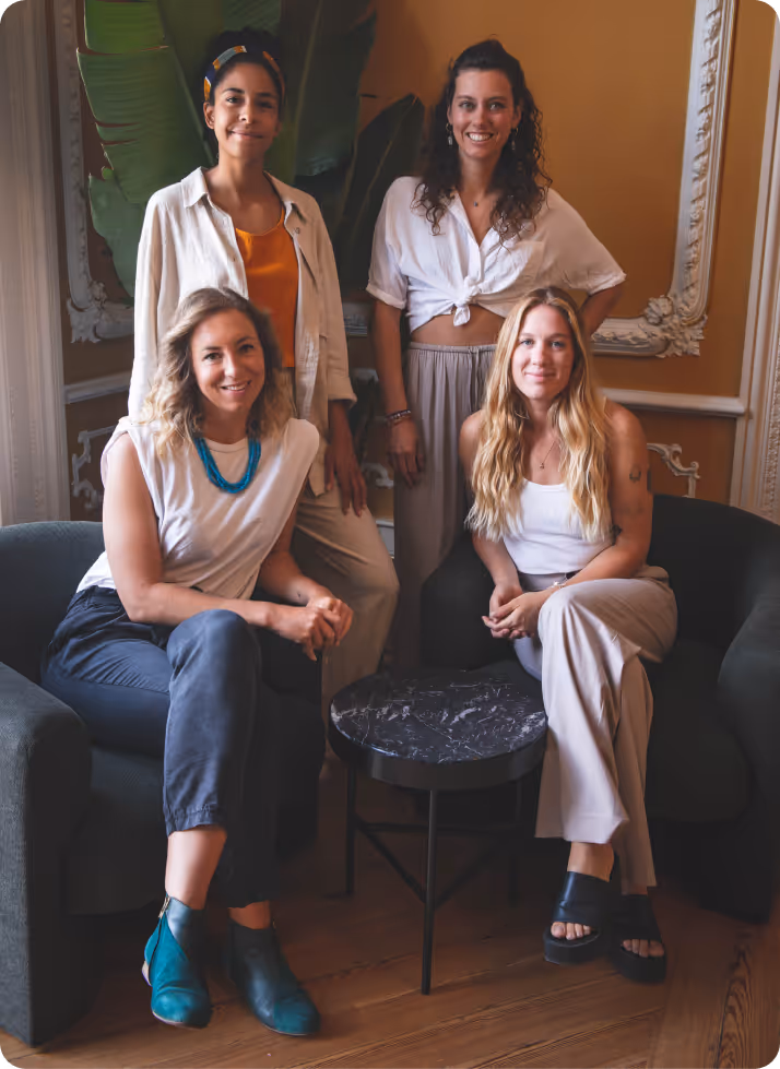Group of four women smiling and posing indoors around a small black round table, two seated on armchairs and two standing behind them.