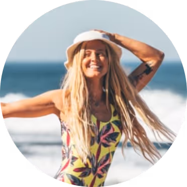 Smiling woman with long blonde hair wearing a white sunhat and colorful swimsuit at the beach with ocean waves in the background.