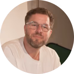 Man with short brown hair, beard, and glasses wearing a white shirt, smiling.