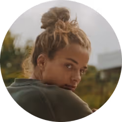 Young woman with light brown hair tied in a messy bun looking over her shoulder outdoors.