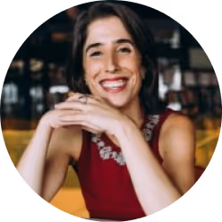 Smiling woman with dark hair wearing a sleeveless red top, seated indoors with blurred background.