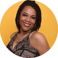 Smiling woman with short dreadlocks wearing a patterned sleeveless top and necklace, against a yellow background.