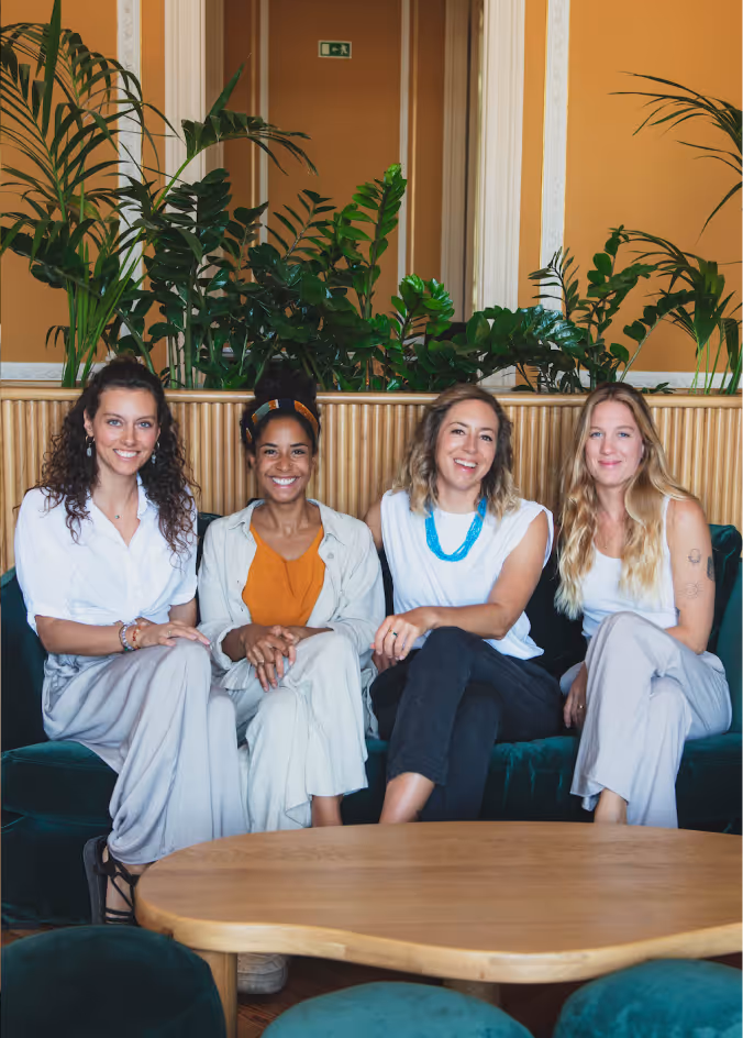 Four women sitting on a green couch in front of a wooden table with plants behind them.
