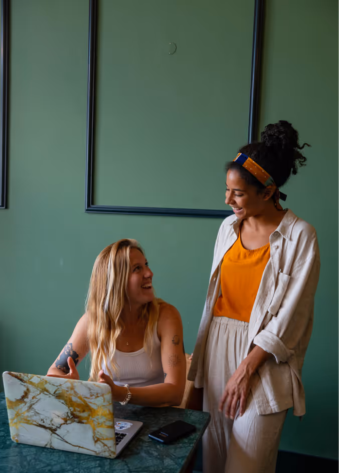 Two women smiling and talking, one seated with a laptop, the other standing against a green wall.