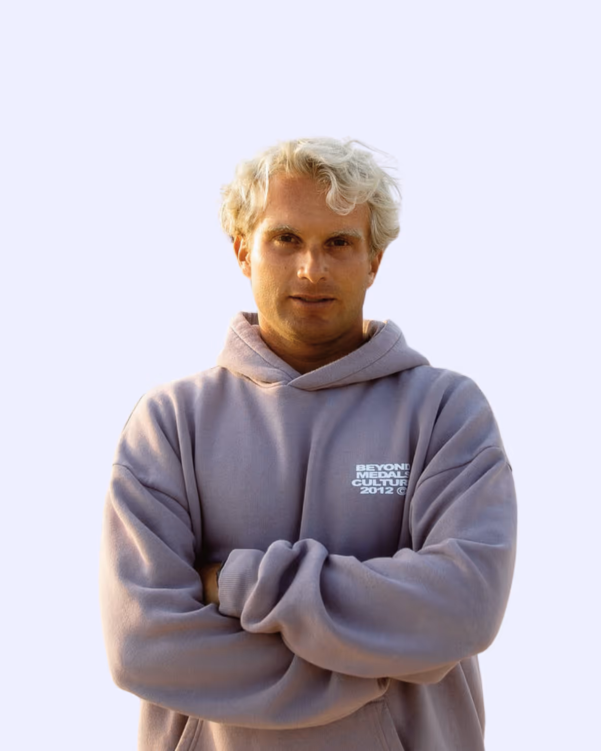 Man with blond curly hair wearing a gray hoodie with text, arms crossed, neutral background.