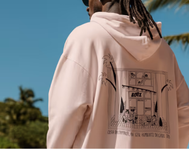Person wearing a light pink hoodie with a black line illustration of a building and palm trees printed on the back.