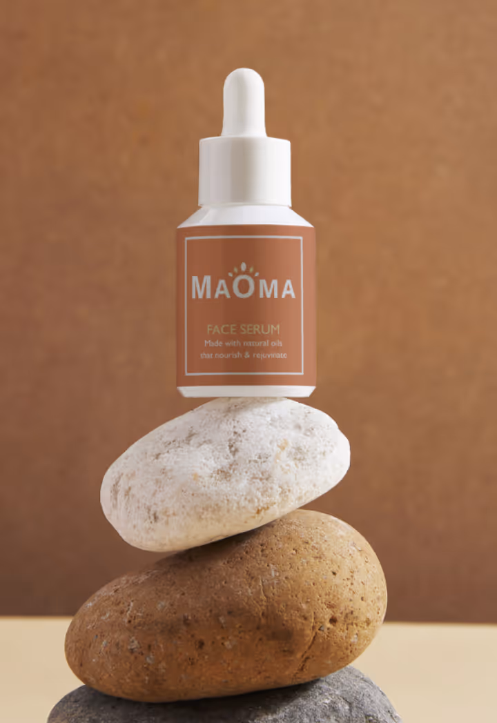 MAOMA face serum bottle with dropper balanced on stacked natural stones against a brown background.