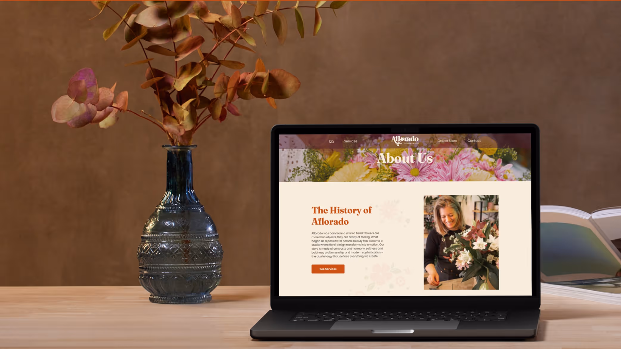 Laptop on wooden table displaying a floral design website's About Us page, with a vase of dried flowers and an open book beside it.