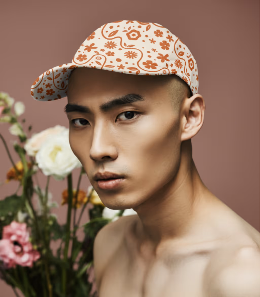 Shirtless young man wearing a white cap with orange floral patterns, standing in front of a blurred background with various flowers.