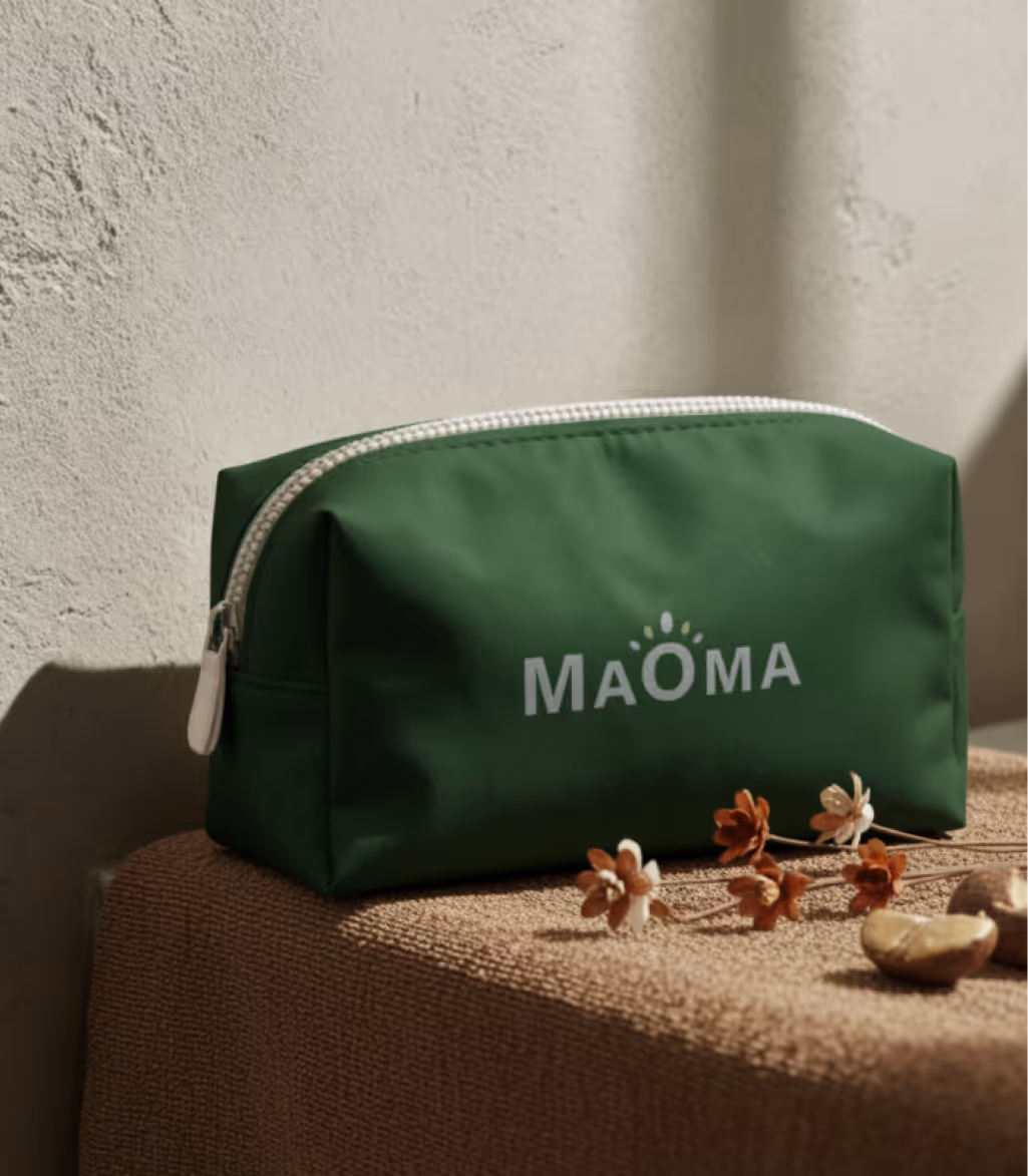 Green rectangular pouch with white zipper and 'MAOMA' printed on front, placed on textured brown surface with dried flowers beside it.