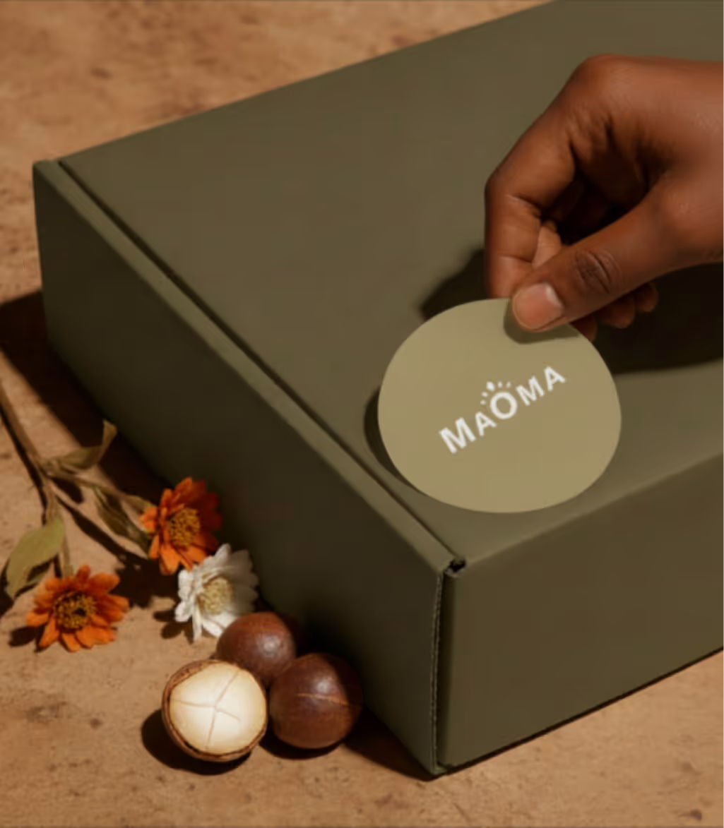 Hand placing a round sticker labeled 'MAÖMA' on an olive green box with flowers and macadamia nuts beside it.