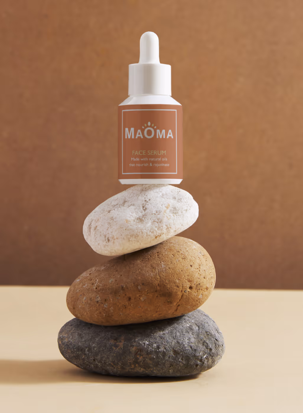 Bottle of Maoma face serum balanced on a stack of three smooth stones against a brown background.