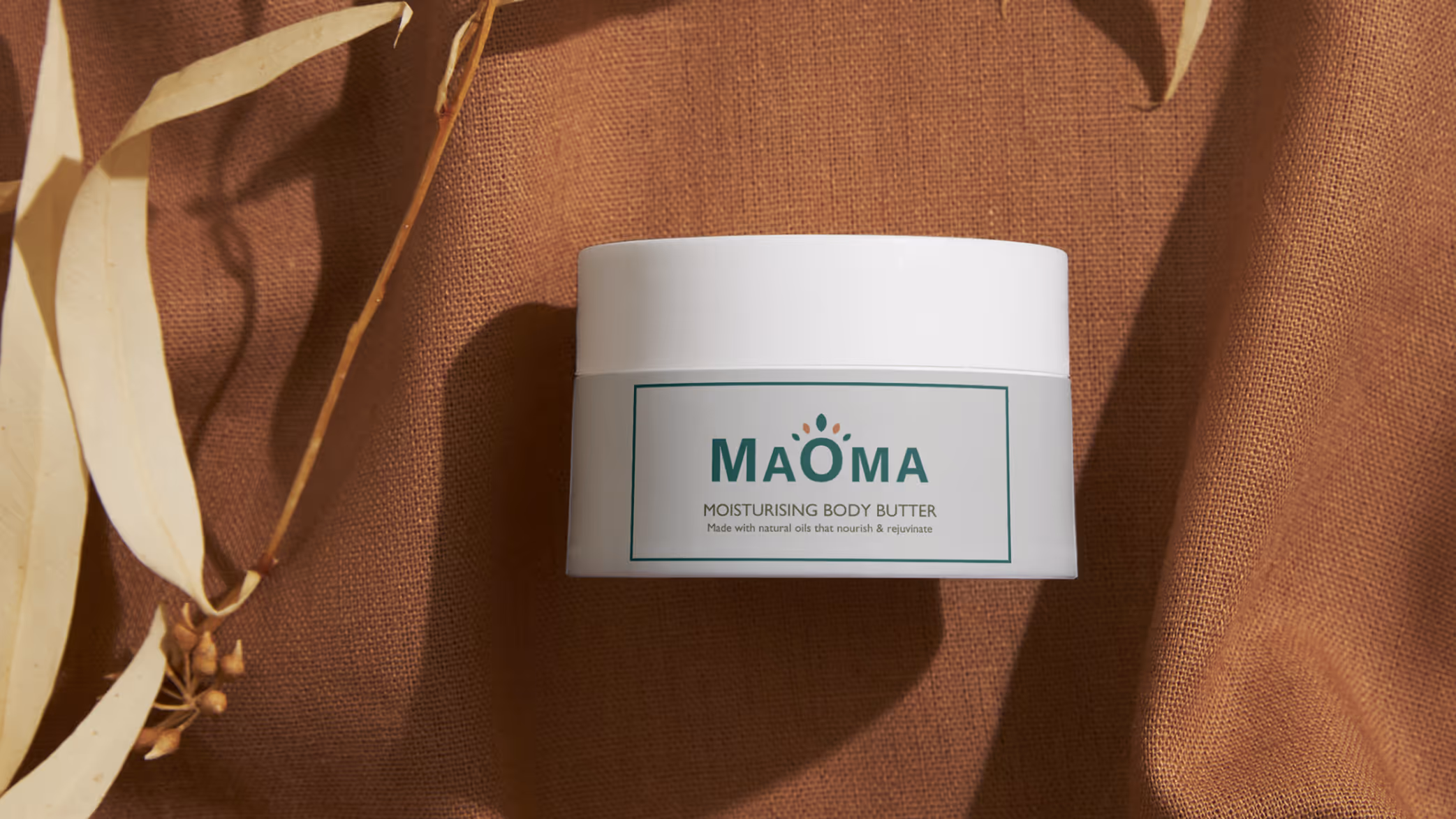 Container of Maoma Moisturising Body Butter on brown textured fabric with dried leaves nearby.