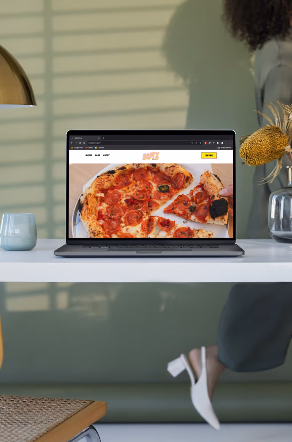 Laptop displaying a pizza ordering website with a pepperoni pizza image, placed on a white desk with a lamp, vase, and a blue cup nearby.