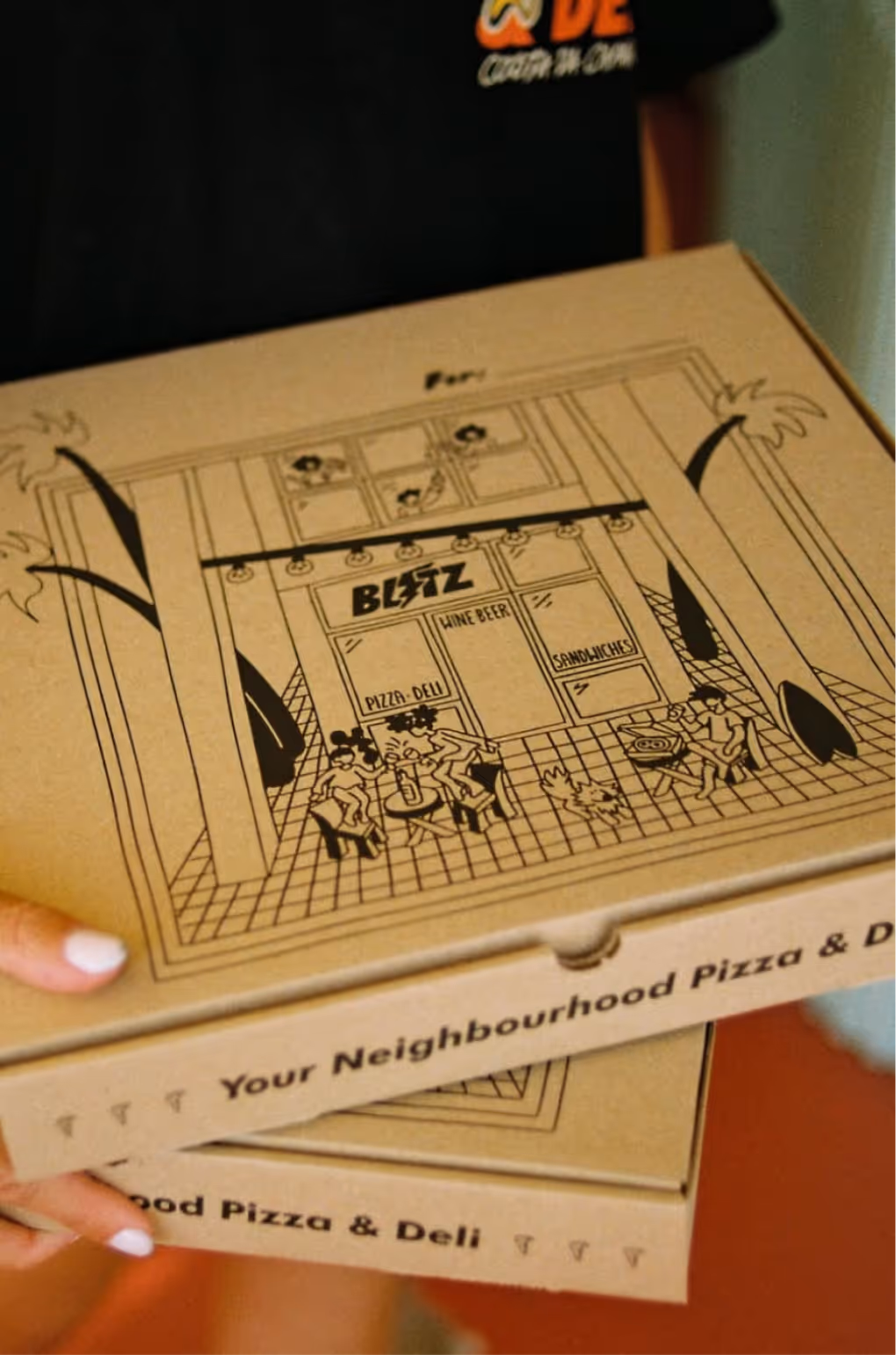 Person holding stacked pizza boxes with a cartoon illustration of a pizzeria named Blitz featuring outdoor tables, palm trees, and people seated.