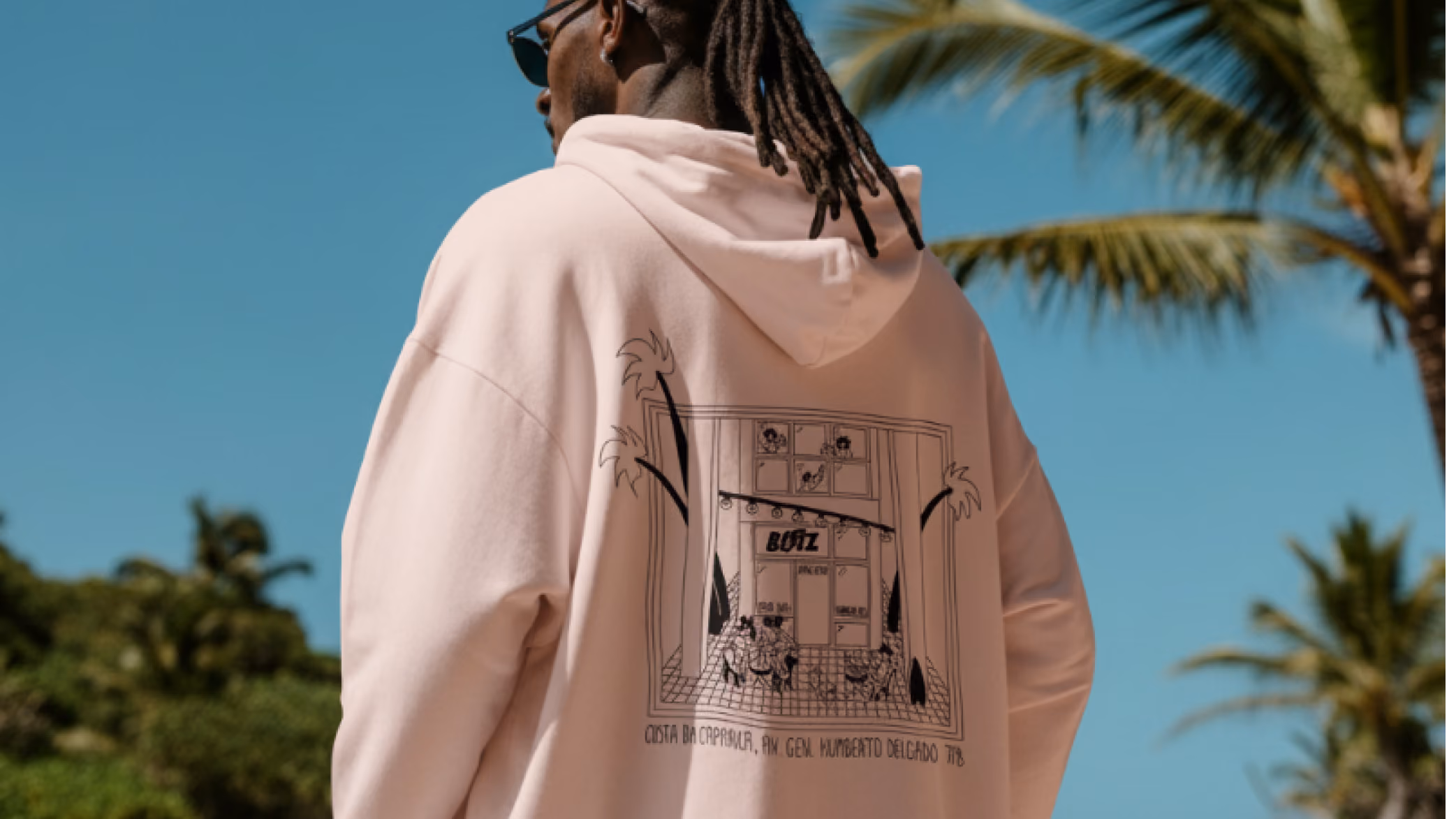 Man wearing a light pink hoodie with a black line drawing of a storefront and palm trees on the back under a clear blue sky.