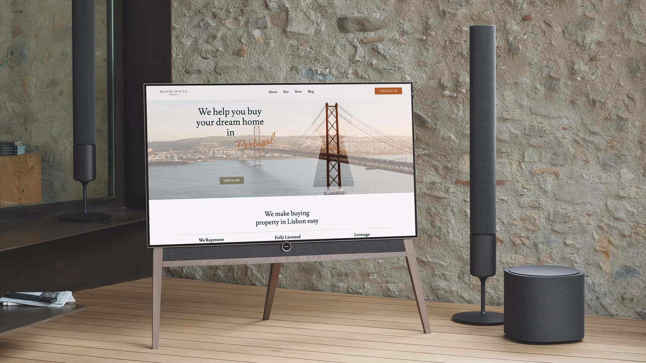 Modern flat-screen TV on wooden stand showing a real estate website with a bridge image and text about buying homes in Portugal, next to black speakers and a stone wall.