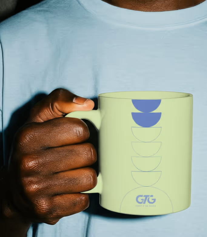 Close-up of a dark-skinned hand holding a light green mug with blue geometric shapes and a GTG logo.
