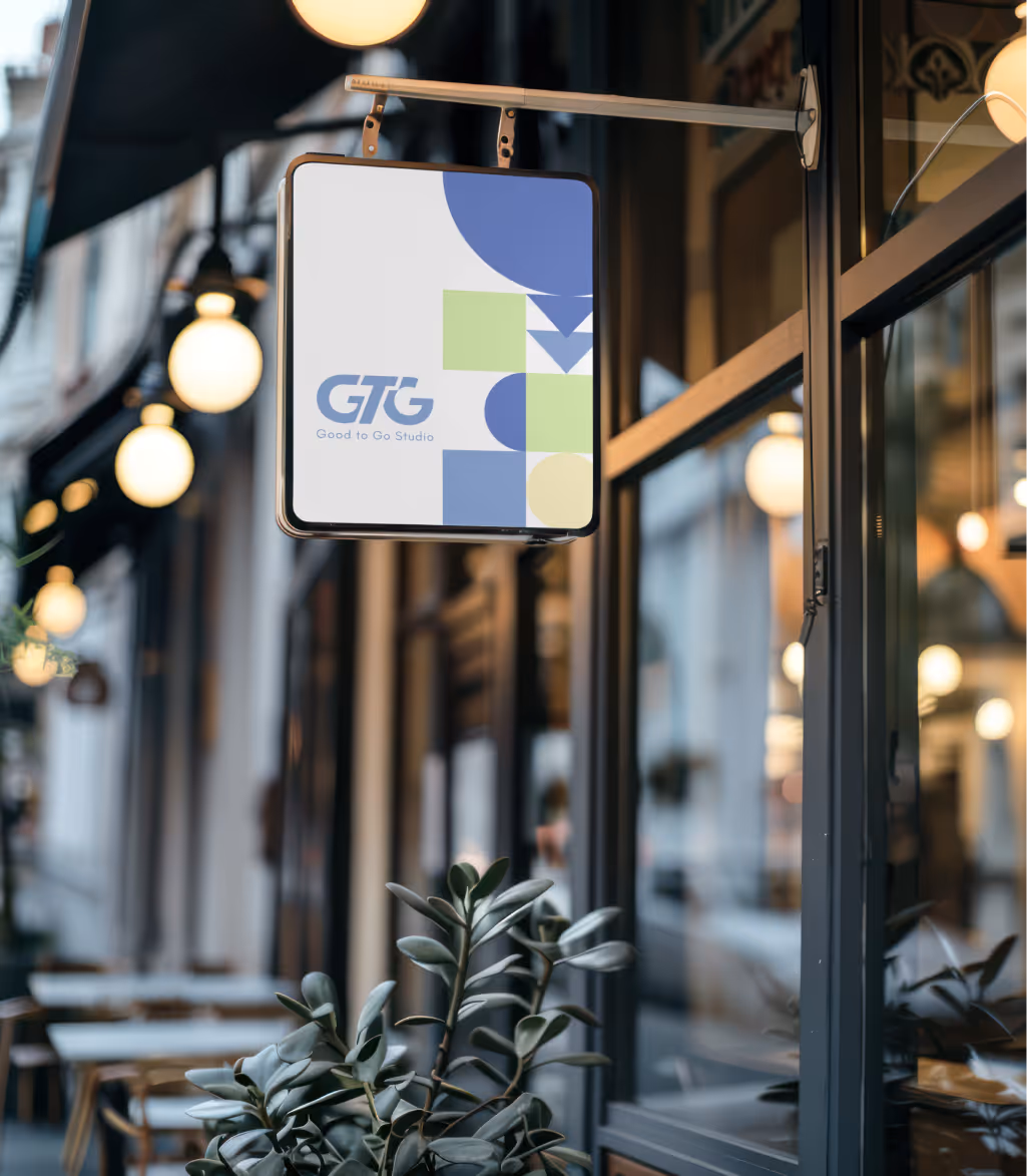 Square hanging sign outside a storefront displaying the GTG Good to Go Studio logo with abstract blue and green shapes.