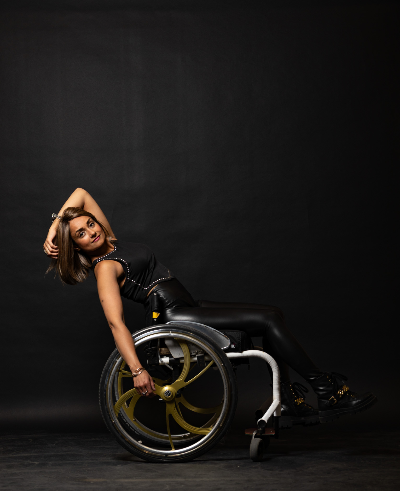 Woman in black outfit posing confidently in a wheelchair against a black background.