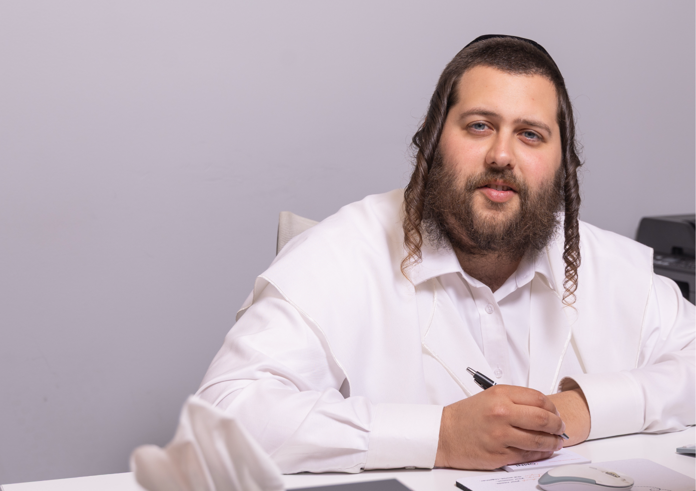 Man with beard and side curls in white shirt writing with pen at desk with tissue box and mouse.