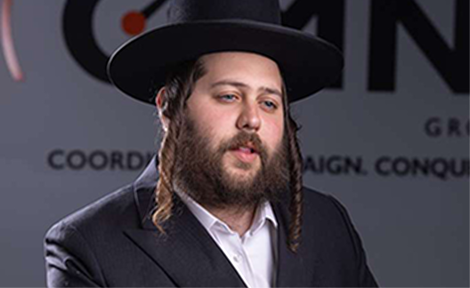 Man with beard and payot wearing a black hat and suit against a blurred sign background.
