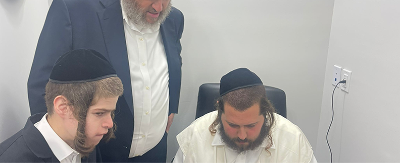 Three men in traditional Jewish attire engaged in reading or study in a small room.