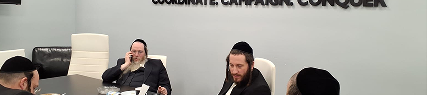 Four men in black hats and suits sitting around a table in a meeting room with a slogan on the wall.