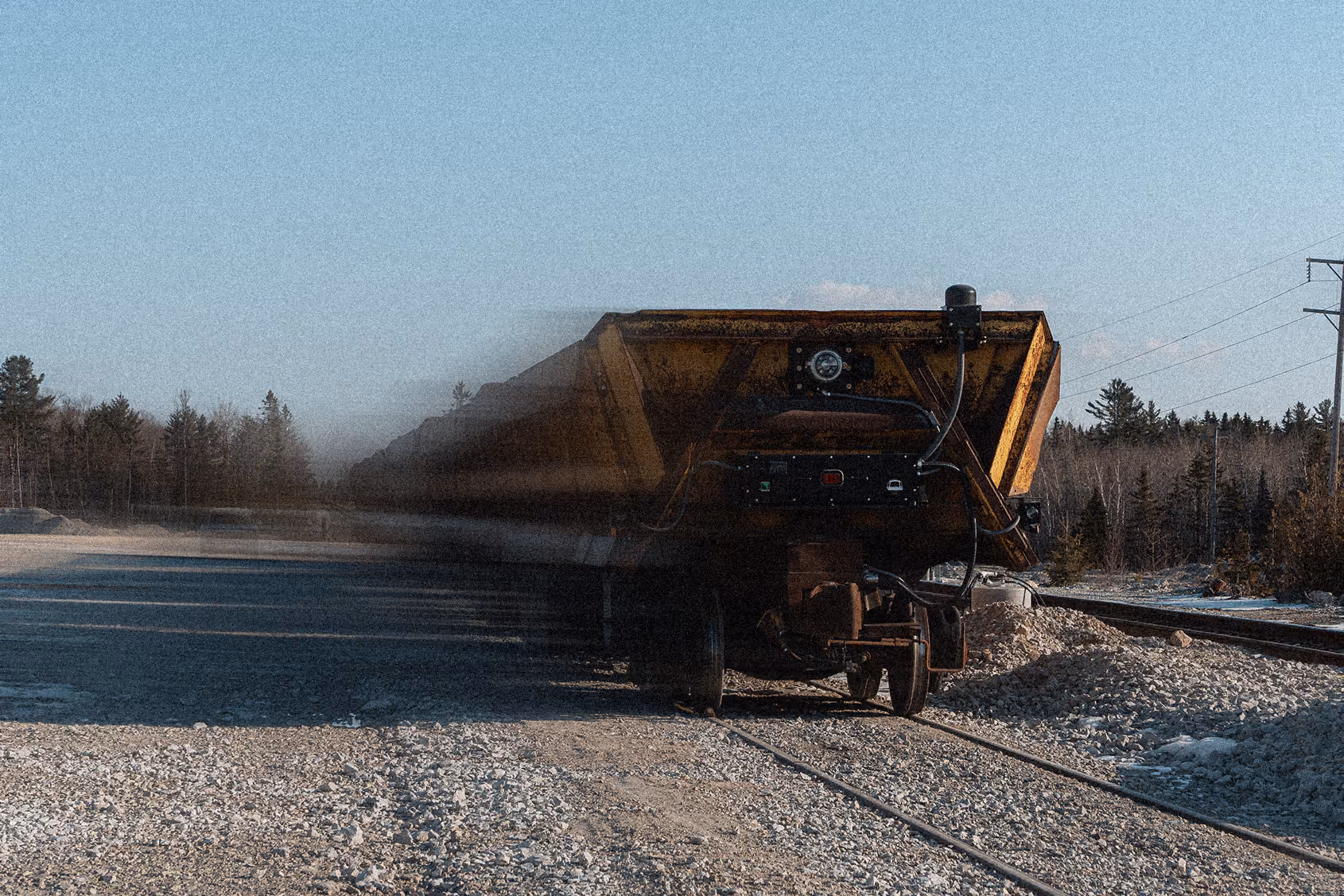 Rusty yellow freight train car on gravel beside railroad tracks with forest and clear blue sky.