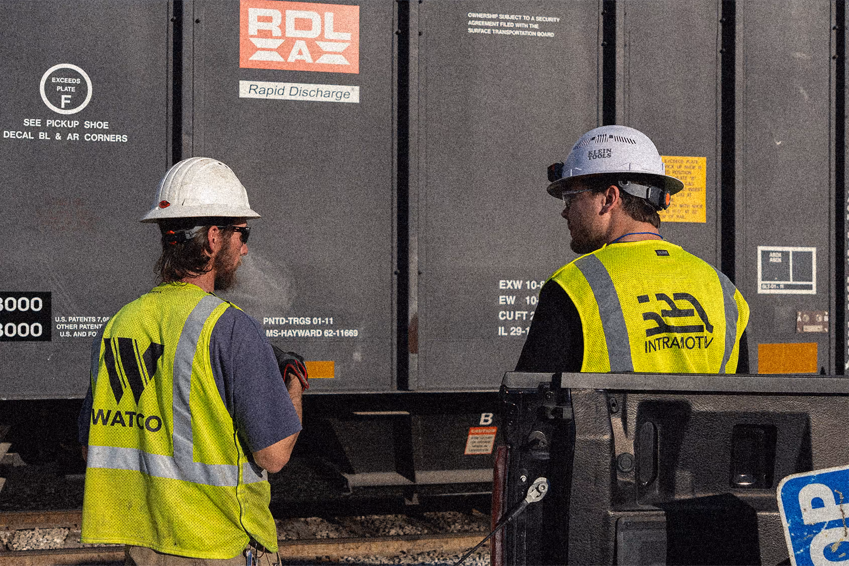 Two construction workers in hard hats and reflective vests talk near a freight train car.