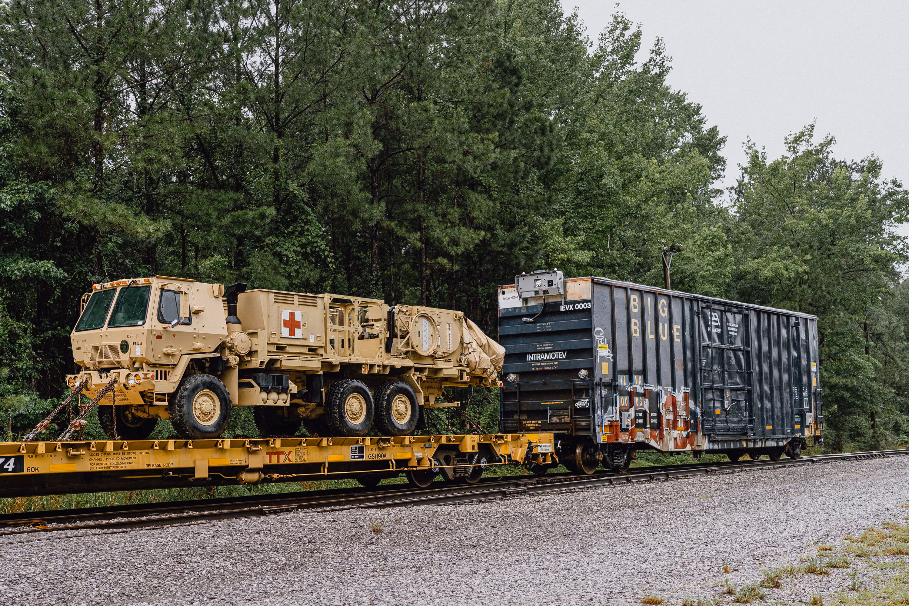 Rail freight car carrying tan military ambulance truck with red cross by green forest.