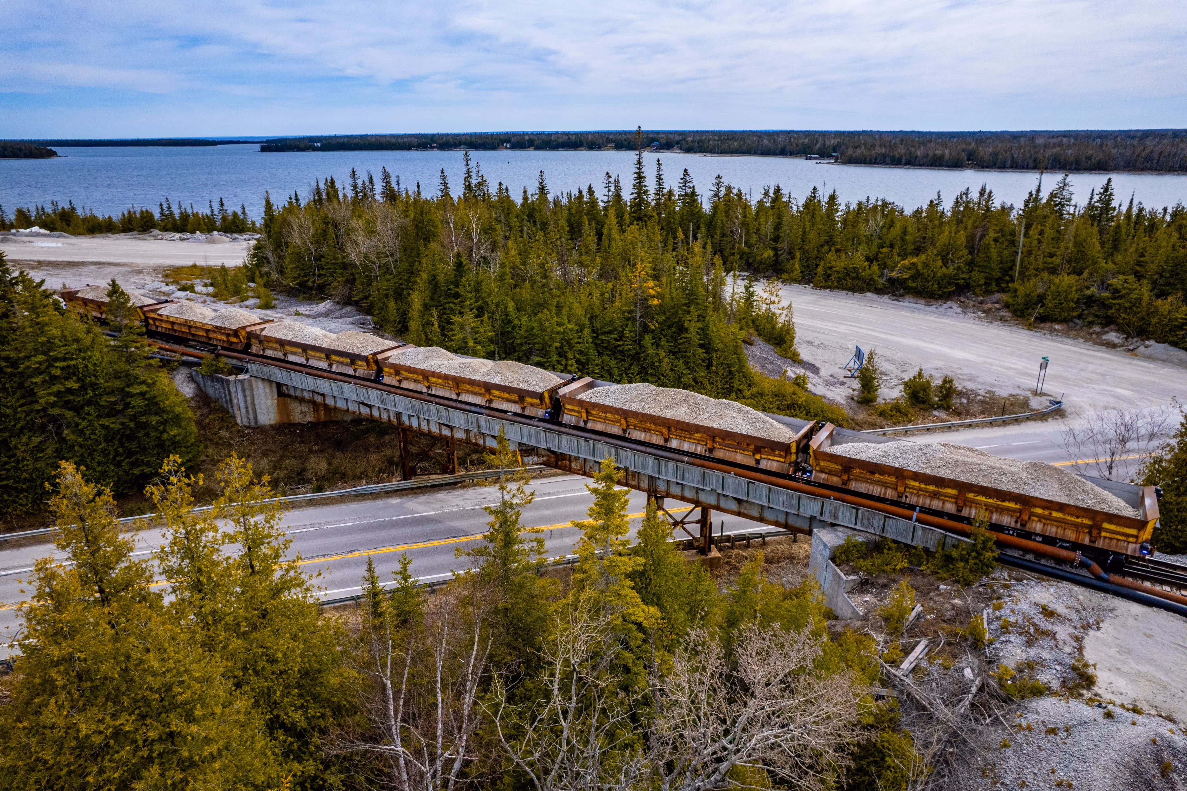 A train carrying gravel crosses a bridge over a road with trees and a lake in the background.