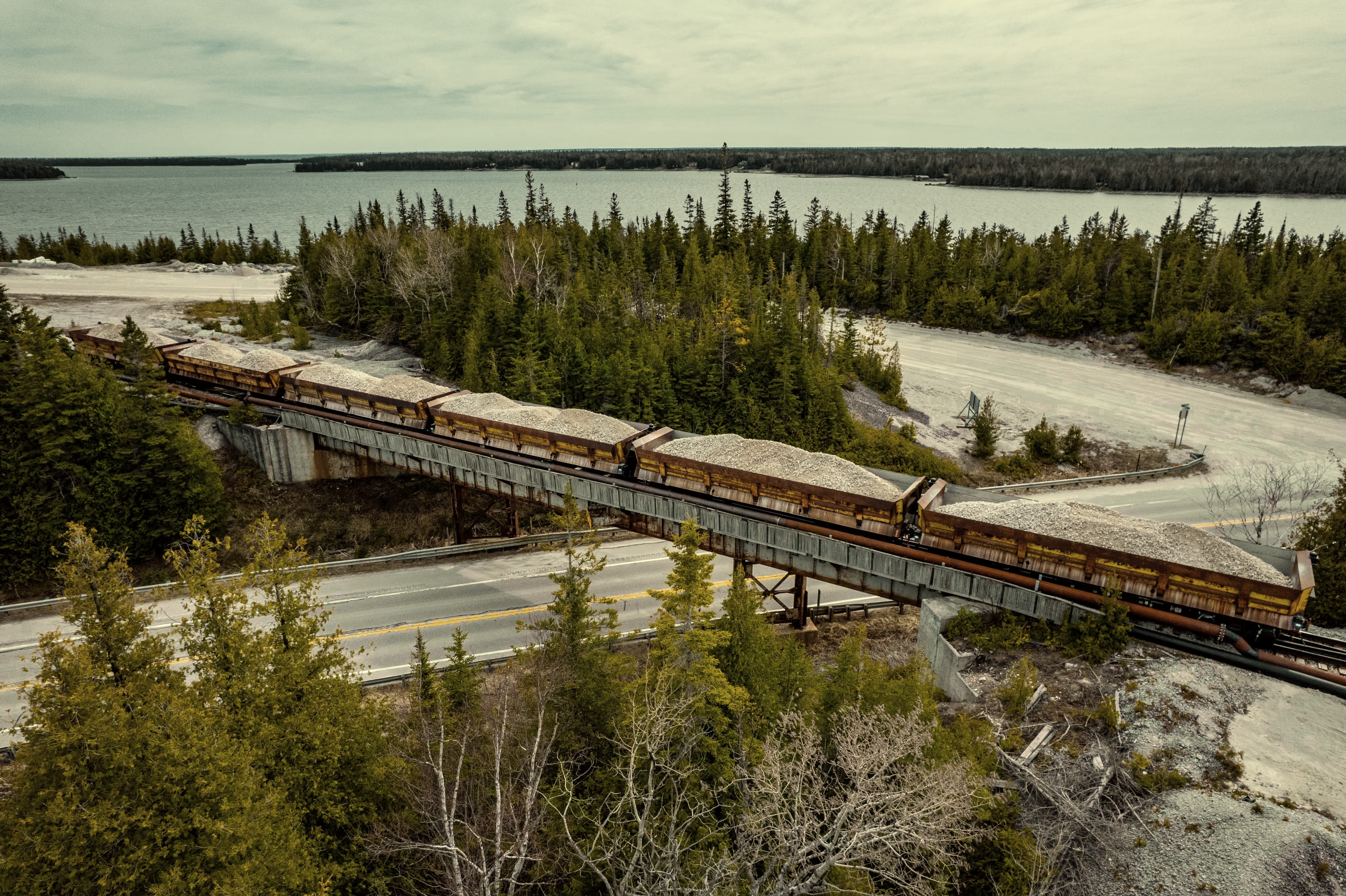 A train carrying gravel crosses a bridge over a road with trees and a lake in the background.