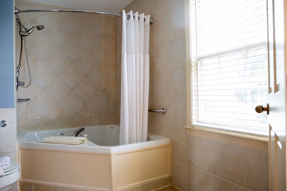 Corner Jacuzzi tub with tiled walls, shower fixture, white curtain, grab bar and large window