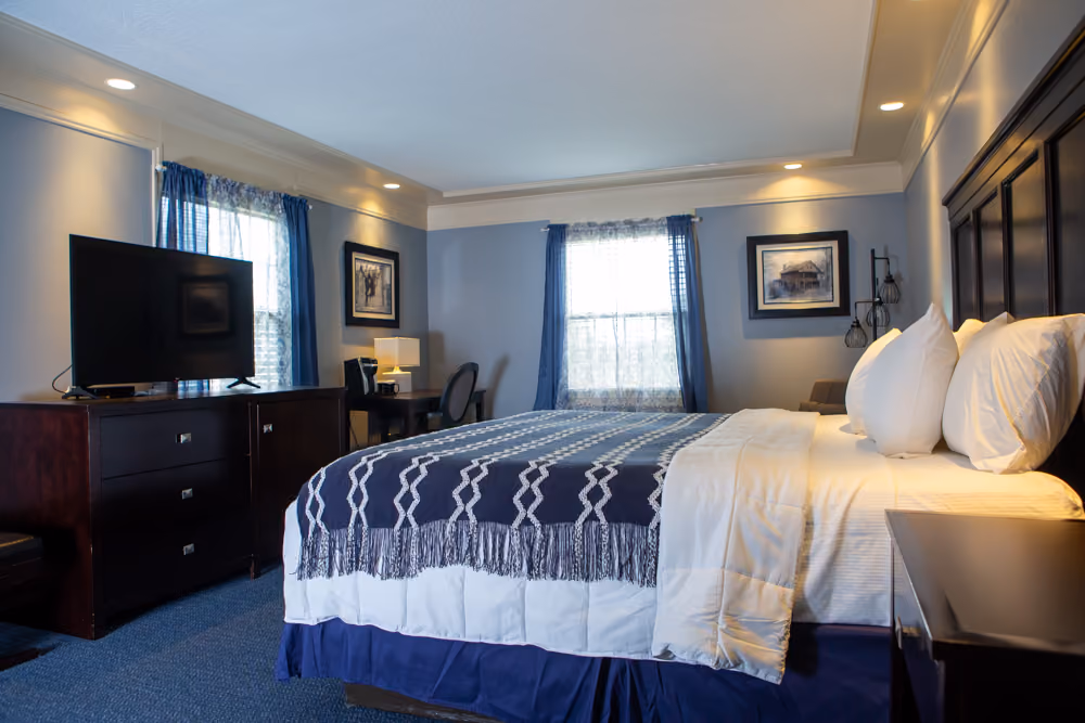 King hotel room with white bedding, navy throw, dark wood furnishings, television, desk and two windows